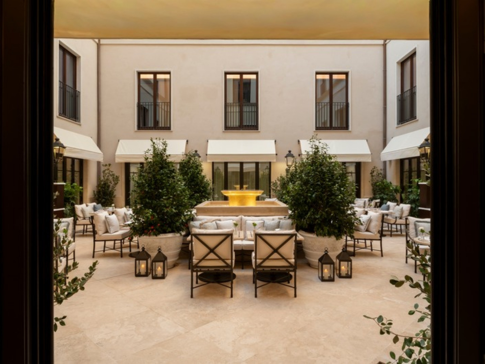 The lantern-lit Piazzetta courtyard at Corinthia Rome, with a central fountain and cushioned armchairs beneath striped awnings